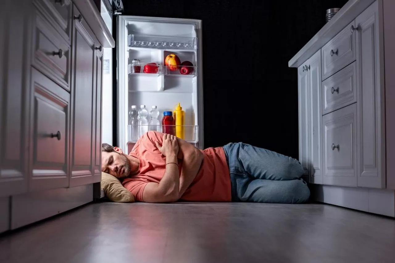 exhausted young man sleeping on kitchen floor near open refrigerator