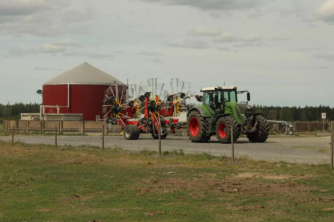 Nowoczesna biogazownia rolnicza w otoczeniu gospodarstwa. Na pierwszym planie ciągnik Fendt z przetrząsaczem/zgrabiarką, a w tle czerwona instalacja biogazowa z kopulastym dachem. Obraz ukazuje potencjał odnawialnych źródeł energii na wsi oraz rolniczą infrastrukturę technologiczną.