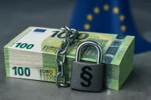 Stos banknotów euro owinięty łańcuchem i zamknięty kłódką z symbolem paragrafu, na tle flagi Unii Europejskiej. Obraz ilustruje kontrowersyjny plan Komisji Europejskiej, by uzależnić dopłaty bezpośrednie dla rolników od oceny praworządności w państwach członkowskich.