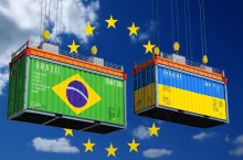 Ilustracja przedstawia dwa kontenery transportowe – zielony z flagą Brazylii i niebiesko-żółty z flagą Ukrainy – unoszące się na tle nieba z gwiazdami symbolizującymi Unię Europejską. Grafika symbolizuje rosnącą presję handlową wywieraną na europejskie rolnictwo w kontekście umów z krajami Mercosur i liberalizacji importu z Ukrainy.