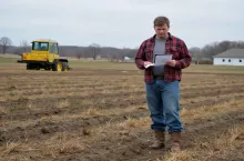 Rolnik stoi z dokumentami na polu, w tle maszyna rolnicza. Zdjęcie symbolizuje niepewność związaną z nowym systemem dopłat unijnych po 2027 roku, które mają być uzależnione od realizacji tzw. kamieni milowych Komisji Europejskiej.