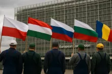 Zdjęcie przedstawia pięciu rolników widzianych od pleców, każdy z flagą swojego kraju – Polski, Węgier, Słowacji, Bułgarii i Rumunii – stojących przed budynkiem Berlaymont w Brukseli. Flagi powiewają na wietrze, a na tle widoczna jest szklana fasada z charakterystycznym niebieskim banerem Komisji Europejskiej. Symboliczna scena protestu rolników z państw przyfrontowych wobec unijnej polityki handlowej.