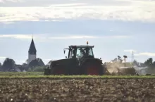 Ciągnik rolniczy wykonujący orkę na polu. W tle widoczna zabudowa wiejska i wieża kościelna. Zdjęcie przedstawia typowy krajobraz rolniczy.