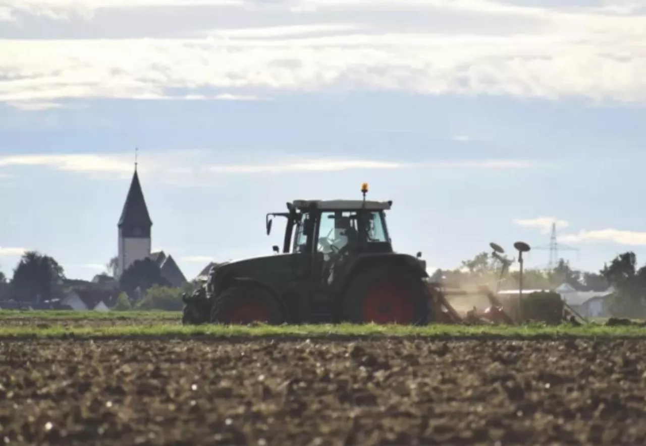 Ciągnik rolniczy wykonujący orkę na polu. W tle widoczna zabudowa wiejska i wieża kościelna. Zdjęcie przedstawia typowy krajobraz rolniczy.