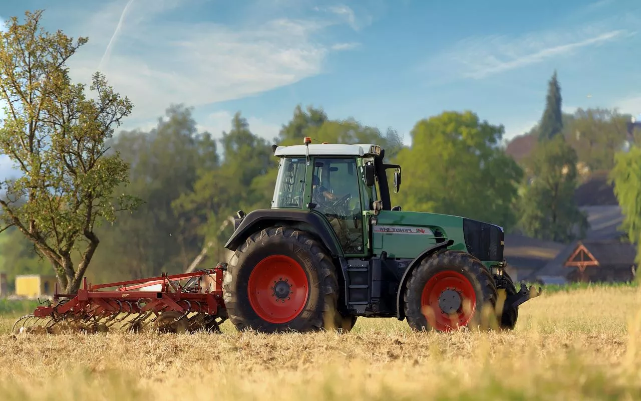 Ciągnik rolniczy marki Fendt podczas pracy na polu, na tle wiejskiego krajobrazu z zabudowaniami i drzewami.