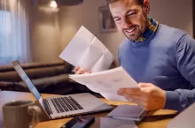 Family budget, paying taxes and bills. A young smiling man holding bills in his hands and calculating payment. He is paying it online on the laptop. Paying bills is easier with e-banking.