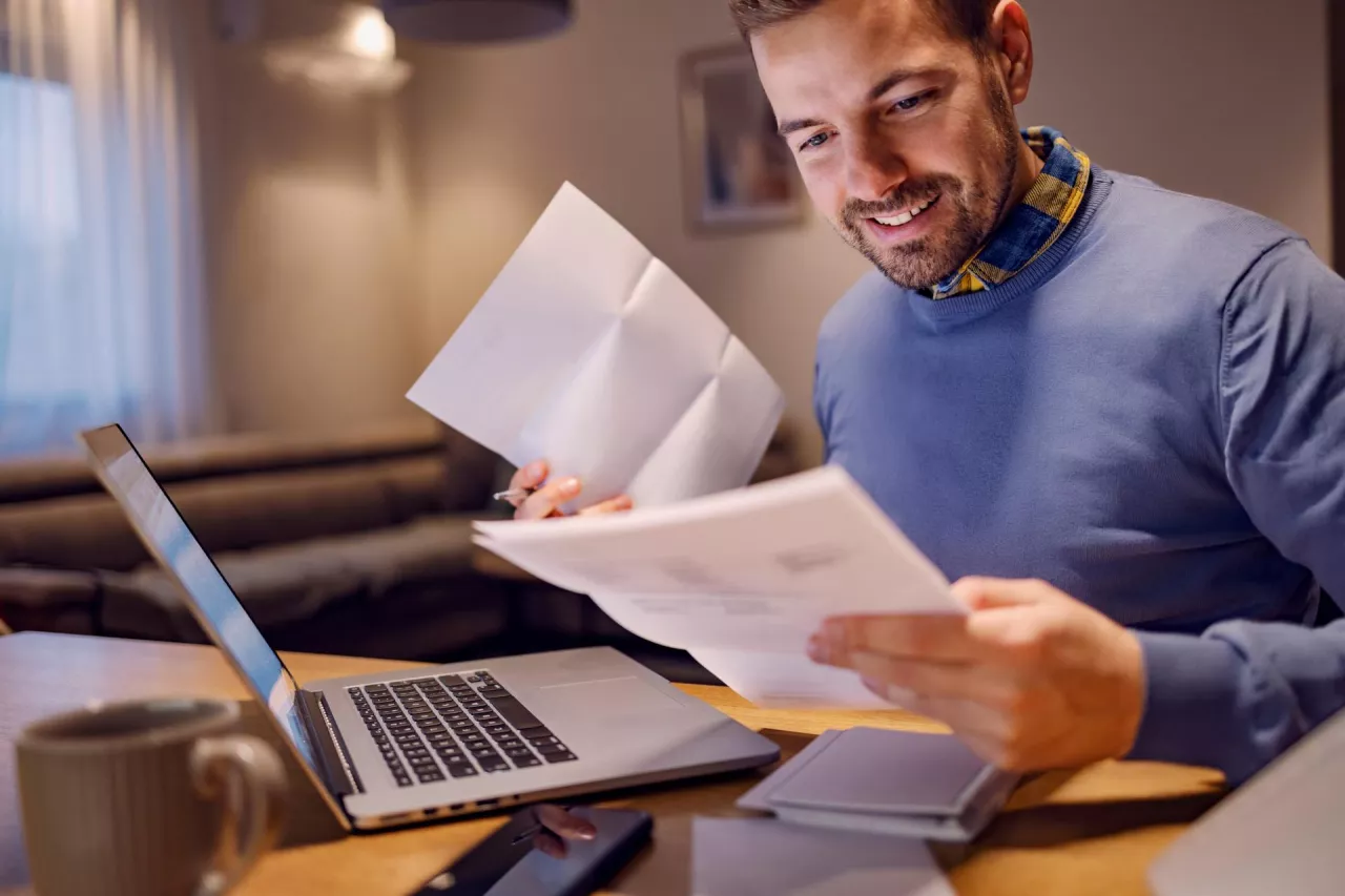 Family budget, paying taxes and bills. A young smiling man holding bills in his hands and calculating payment. He is paying it online on the laptop. Paying bills is easier with e-banking.