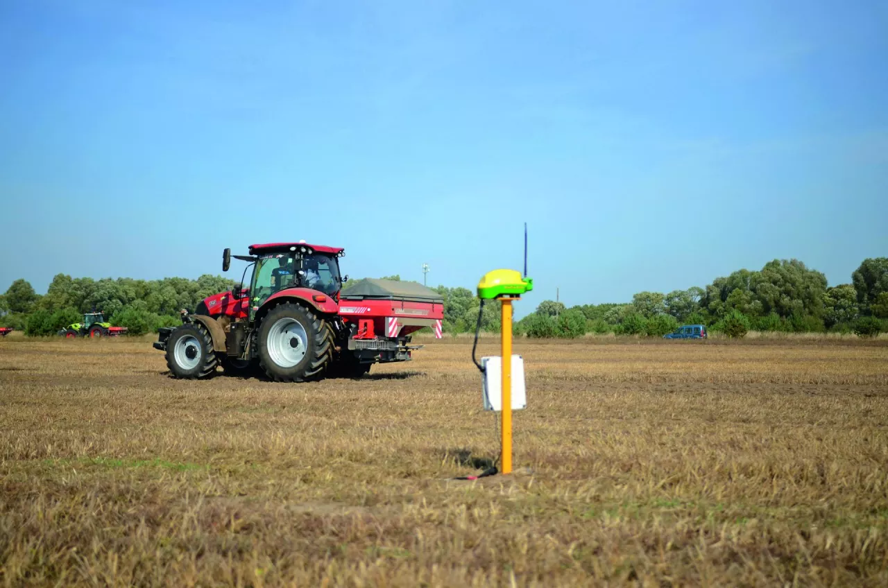 Ciągnik z zaczepionym rozsiewaczem nawozów jedzie na polu, na pierwszym planie widoczna jest antena GPS