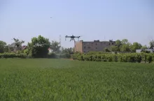 Dron latający nad polem. W tle widoczne bloki mieszkalne