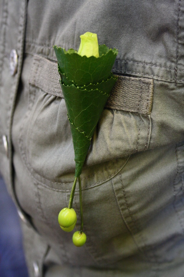 Przypinka dla chłopca wykonana ze zwiniętych liści, białej różyczki i zwieszających się owoców dziurawca ozdobnego. Mocowanie do butonierki drutem owiniętym zieloną taśmą florystyczną.